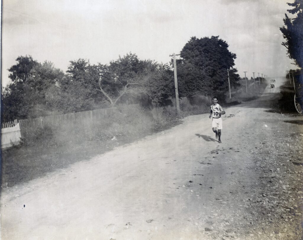 Sam Mellor runs in his race jersey and shorts along a dusty road during the 1904 Olympic marathon, a historical black-and-white photograph.